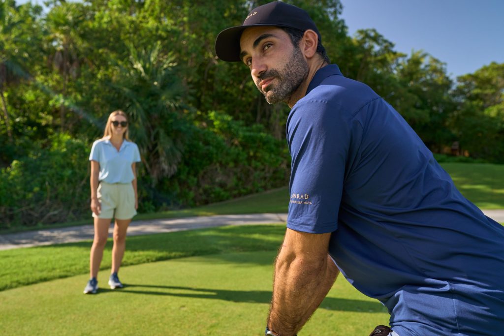 A man in blue tee and black cap plays golf and lady watches nearby.