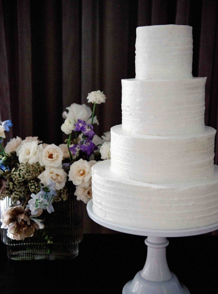 Una tarta nupcial de cuatro pisos se alza elegante sobre la mesa junto a un ramo de flores.