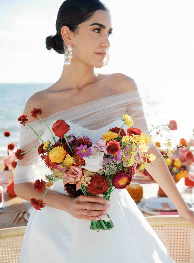 Una dama vestida de novia se encuentra cerca de la mesa del comedor de la playa, sosteniendo un ramo.