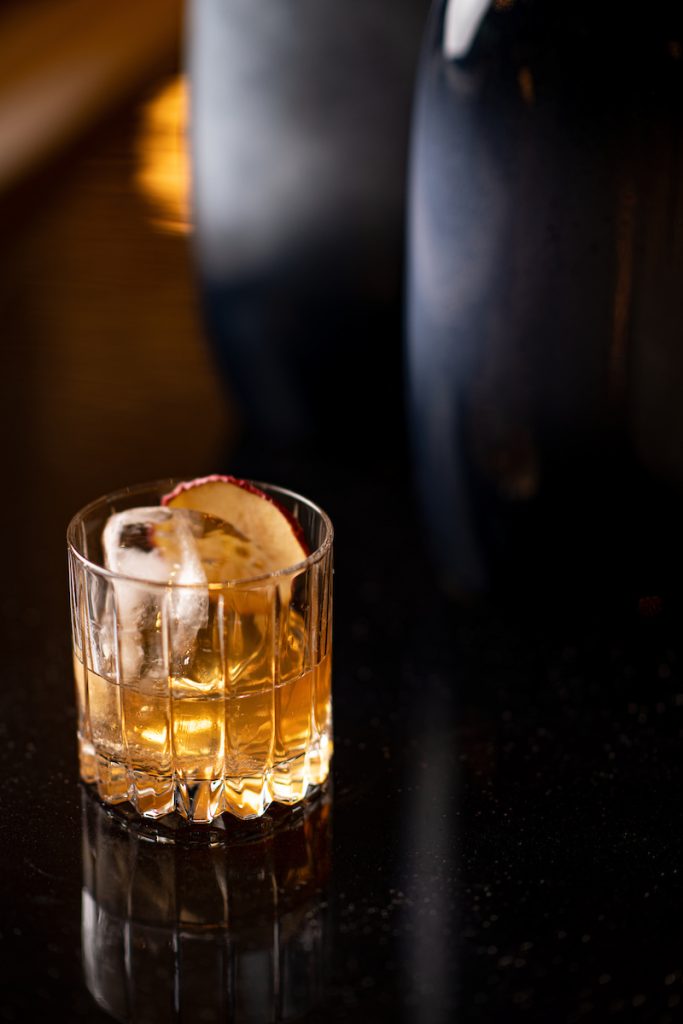 Un vaso de whisky medio lleno descansa sobre la mesa, con el líquido ámbar brillando suavemente.