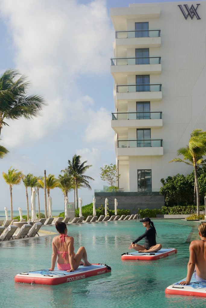 Guests practicing yoga on floating mats in the pool beside the resort tower and palms