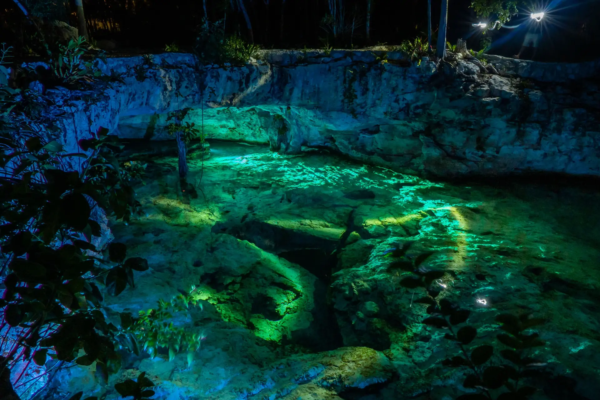 Cenote iluminado de noche con iluminación subacuática turquesa y verde sobre roca caliza en la Riviera Maya cerca de Cancún